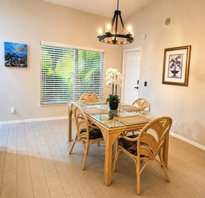 A dining area with a glass table, wicker chairs, a chandelier, and artwork on the walls.