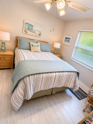 A bedroom with a bed, a ceiling fan, two bedside tables, a window with blinds, and wall art.