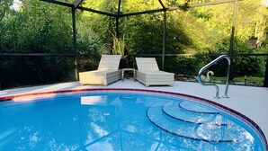 Une piscine avec deux chaises longues et une petite table, le tout entouré d’une structure en verre offrant une vue sur la verdure.