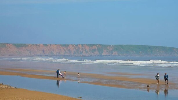 A beach with people walking and playing, a calm sea, and a cliff in the background.