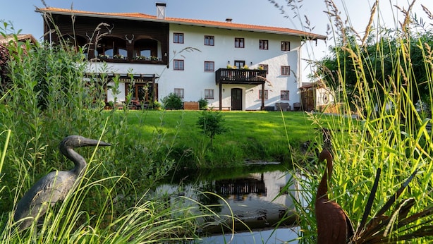 Um edifício de dois andares com uma varanda, um lago ao primeiro plano e vegetação ao redor.