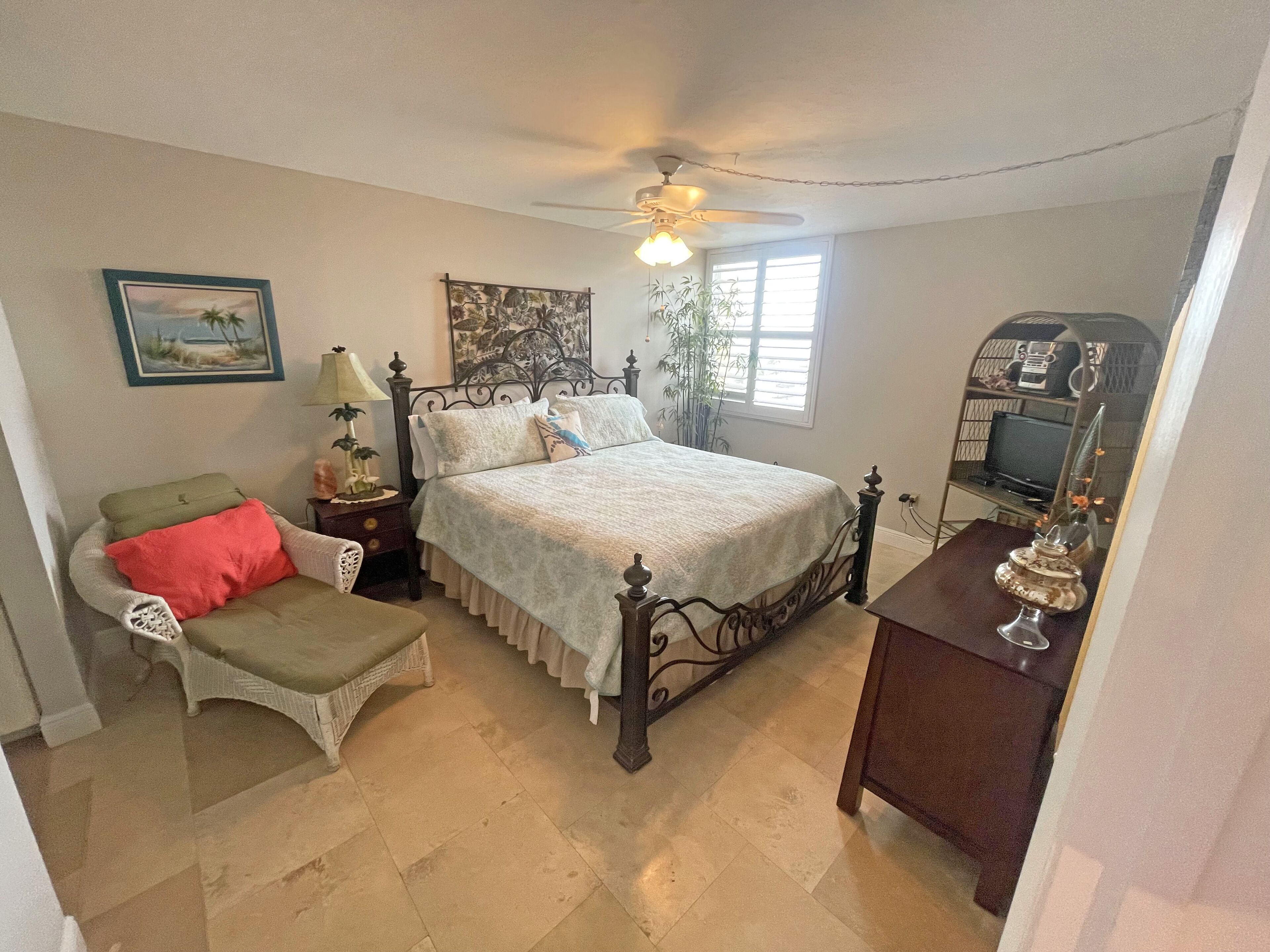 A bedroom with a bed, a chair, a bedside table, a ceiling fan, a window, and a television.