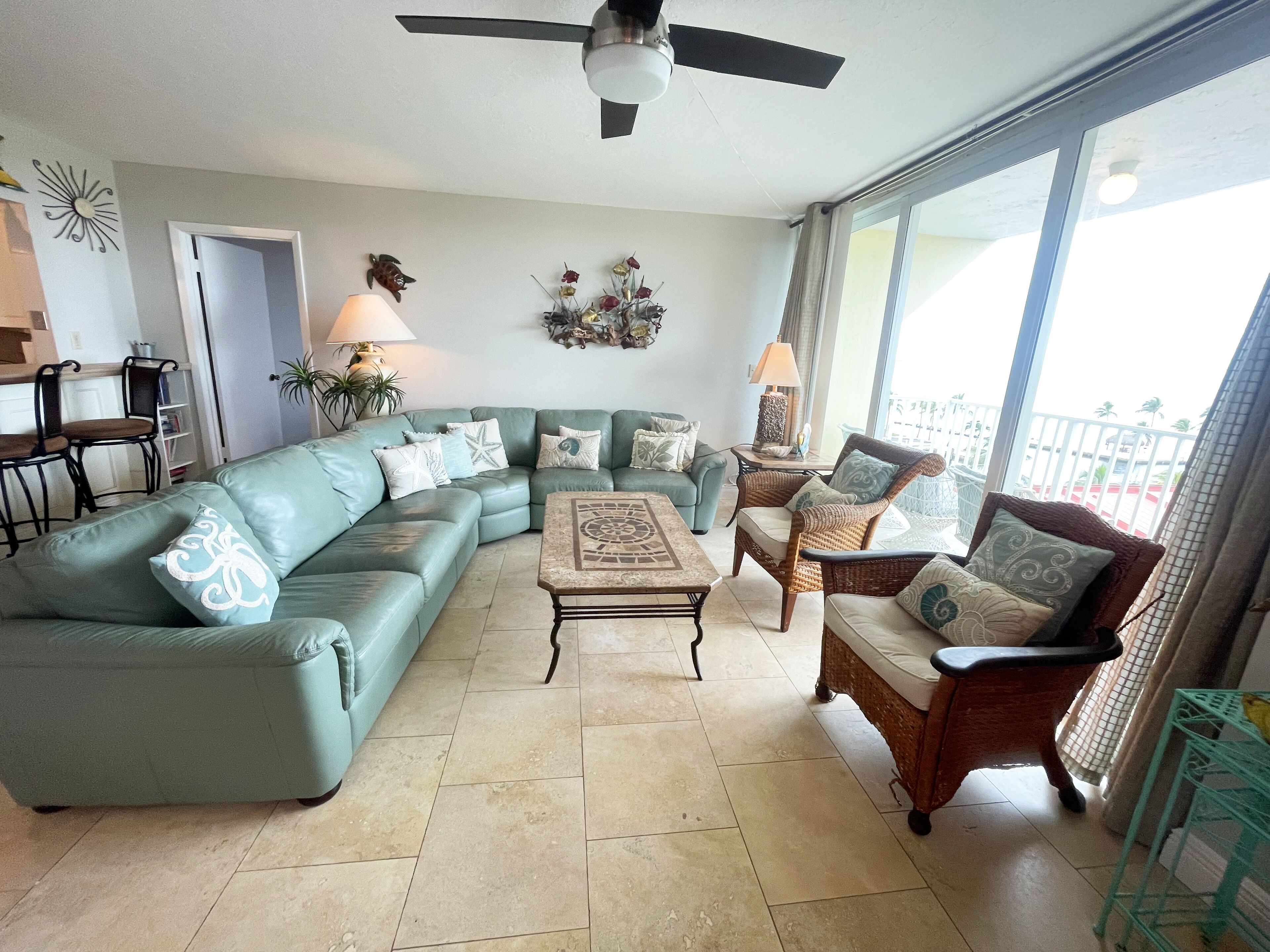 A living room with a sectional sofa, a coffee table, and two wicker chairs. There is a ceiling fan, a window with a view, and a door leading to another room.
