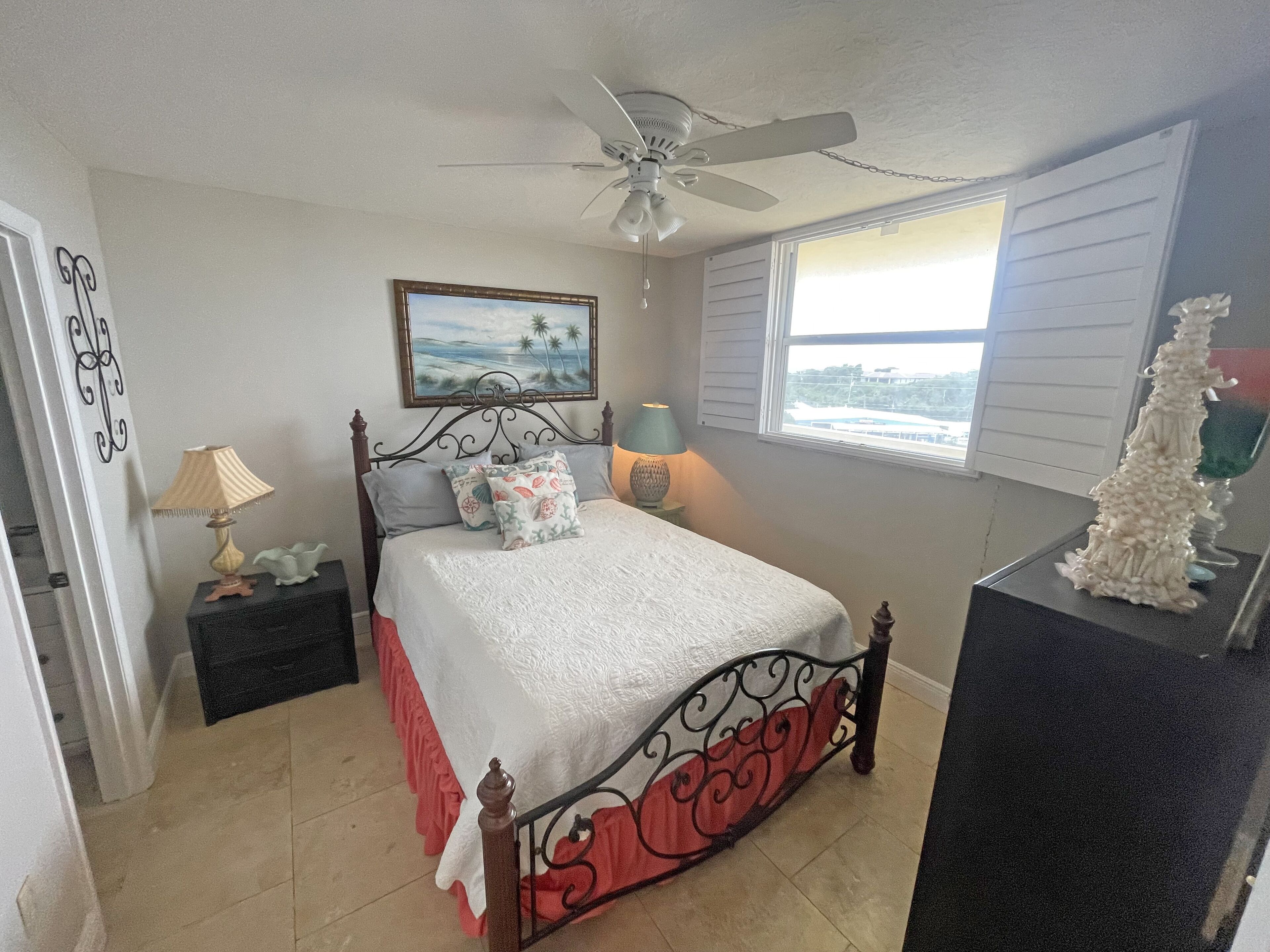 A bedroom with a bed, a ceiling fan, a window with blinds, a nightstand with a lamp, and a decorative item on a dresser.
