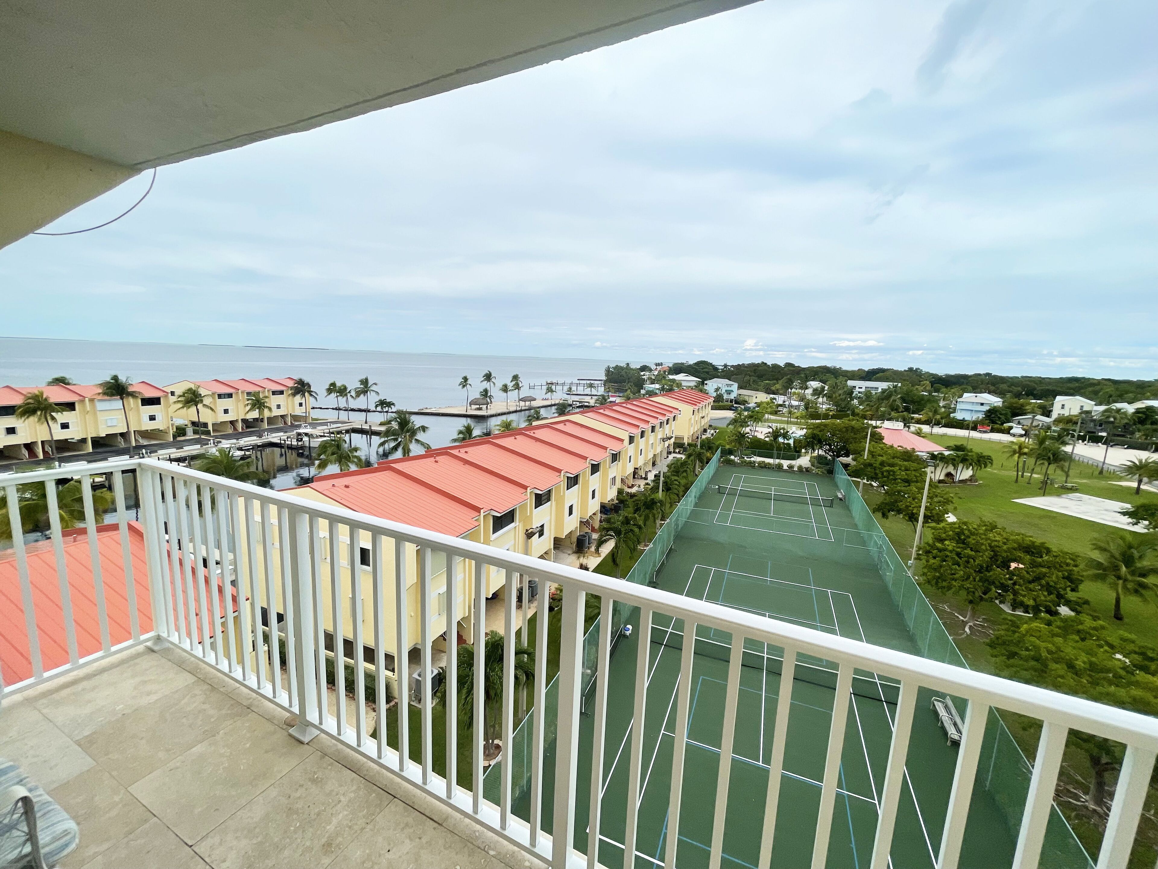 A balcony with a view of a tennis court, a pool, and a row of buildings with red roofs.
