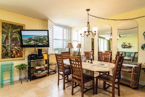 Un comedor con una mesa grande, varias sillas, un televisor y un cuadro de una escena tropical.