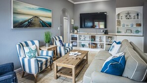 A living room with striped chairs, a wooden coffee table, a grey sofa with blue pillows, a flat-screen TV, and a shelf with decorative items.