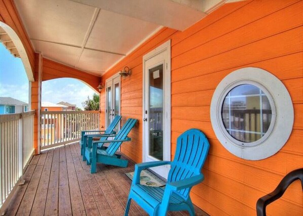 A covered porch with wooden flooring and a railing, featuring blue Adirondack chairs and a round window on the wall.