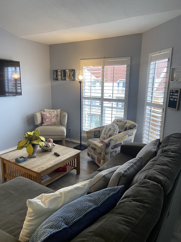 A living room with a sofa, armchair, and a coffee table. There is a window with blinds, a lamp, and a TV mounted on the wall. - Condo on the Beach! Ocean view, pool, hot tub, sauna, gym and nearby restaurants (Corpus Christi)