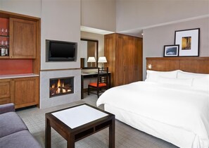 A hotel room with a large bed, a TV mounted on the wall, a fireplace, a desk with a chair, and wooden cabinets.