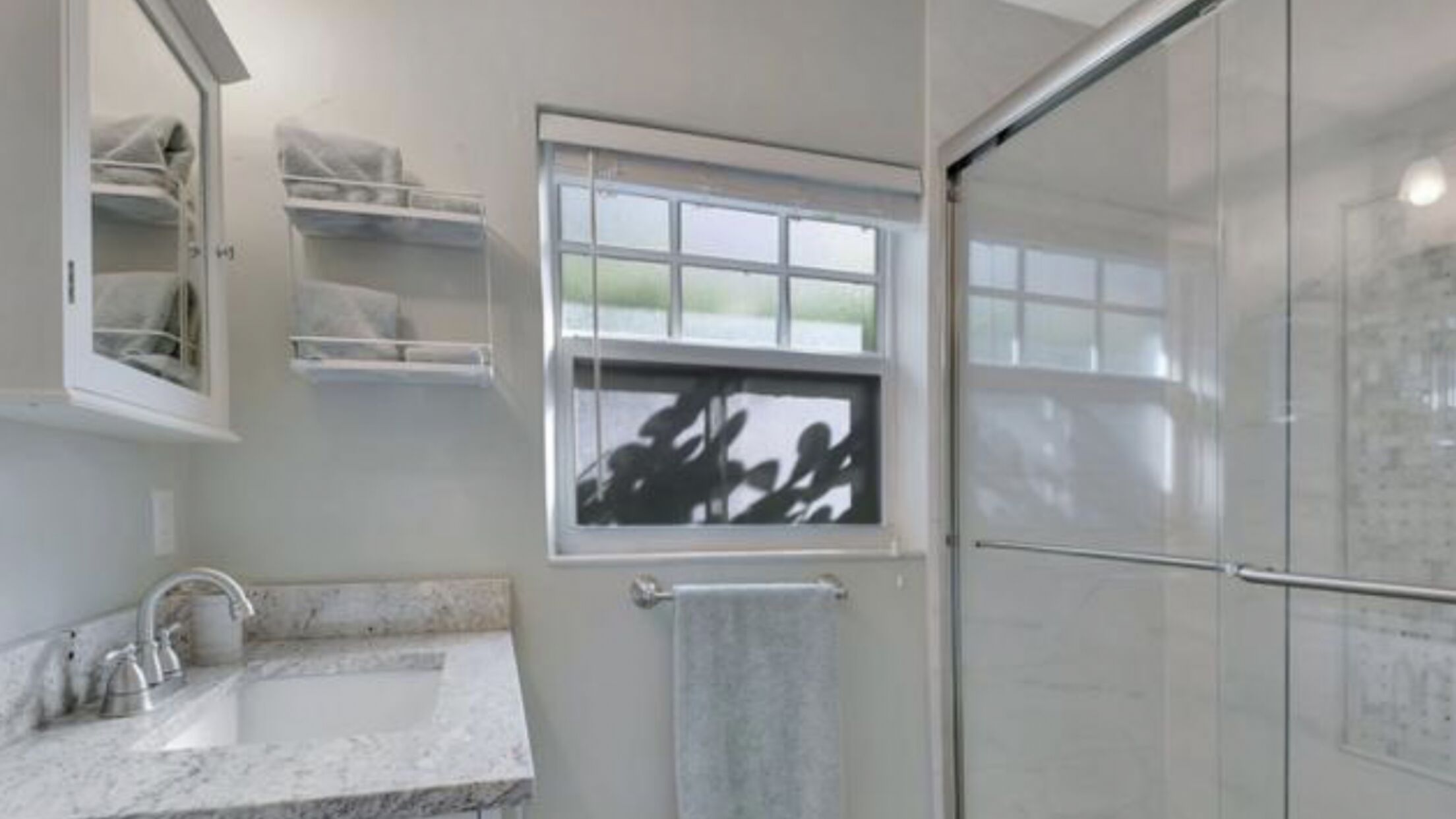 A bathroom with a shower, a sink with a marble countertop, a towel rack, and a window with a view of trees.