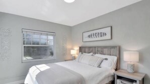 A bedroom with a large bed, two bedside tables with lamps, a window, and a wall-mounted artwork.