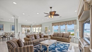A spacious living room with a large window overlooking the ocean, furnished with a patterned sofa, a coffee table, and a ceiling fan. - Pineapple Palace, Luxury Oceanfront Beach House with Pool and Hot Tub (North Myrtle Beach)