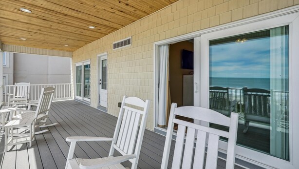 Une terrasse extérieure couverte, dotée de chaises blanches et d’une porte vitrée donnant sur un balcon avec vue sur l’océan.