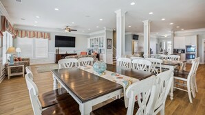 Un salon spacieux avec une grande table à manger en bois, des chaises blanches et un téléviseur fixé au mur.