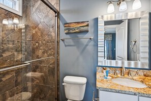 A bathroom with a glass shower enclosure, a toilet, a sink with a marble countertop, and a large mirror with a landscape painting above it. - Sun Sand & Sea, an Oceanfront 9BR House with Private Pool. (North Myrtle Beach)