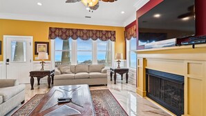 A living room with a beige sofa, a coffee table, and a fireplace. There is a large window with a view of the ocean. - Sun Sand & Sea, an Oceanfront 9BR House with Private Pool. (North Myrtle Beach)