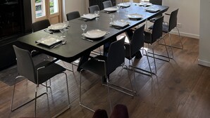 Un comedor moderno con una larga mesa dispuesta para seis personas, rodeada de sillas negras, ventanas amplias y un techo con vigas a la vista.