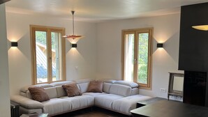 Una sala de estar moderna con un sofá modular, un sillón oscuro y una mesa de comedor. Hay dos ventanas con vistas al exterior e iluminación empotrada.