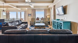 A spacious living room with a black leather sofa, a dining area with a table and chairs, a ceiling fan, and a TV mounted on the wall. - Living Beach 2, Luxury Oceanfront Home in Cherry Grove with Game Room and Pool (North Myrtle Beach)