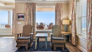 A balcony with two wooden chairs, a small table, and a lamp. There is a framed picture on the wall and a window with curtains. - Living Beach 2, Luxury Oceanfront Home in Cherry Grove with Game Room and Pool (North Myrtle Beach)