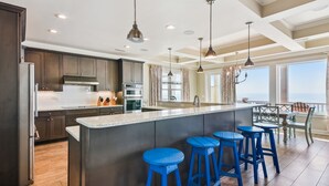 A modern kitchen with dark wood cabinets, stainless steel appliances, and a large island with blue bar stools. - Living Beach 2, Luxury Oceanfront Home in Cherry Grove with Game Room and Pool (North Myrtle Beach)