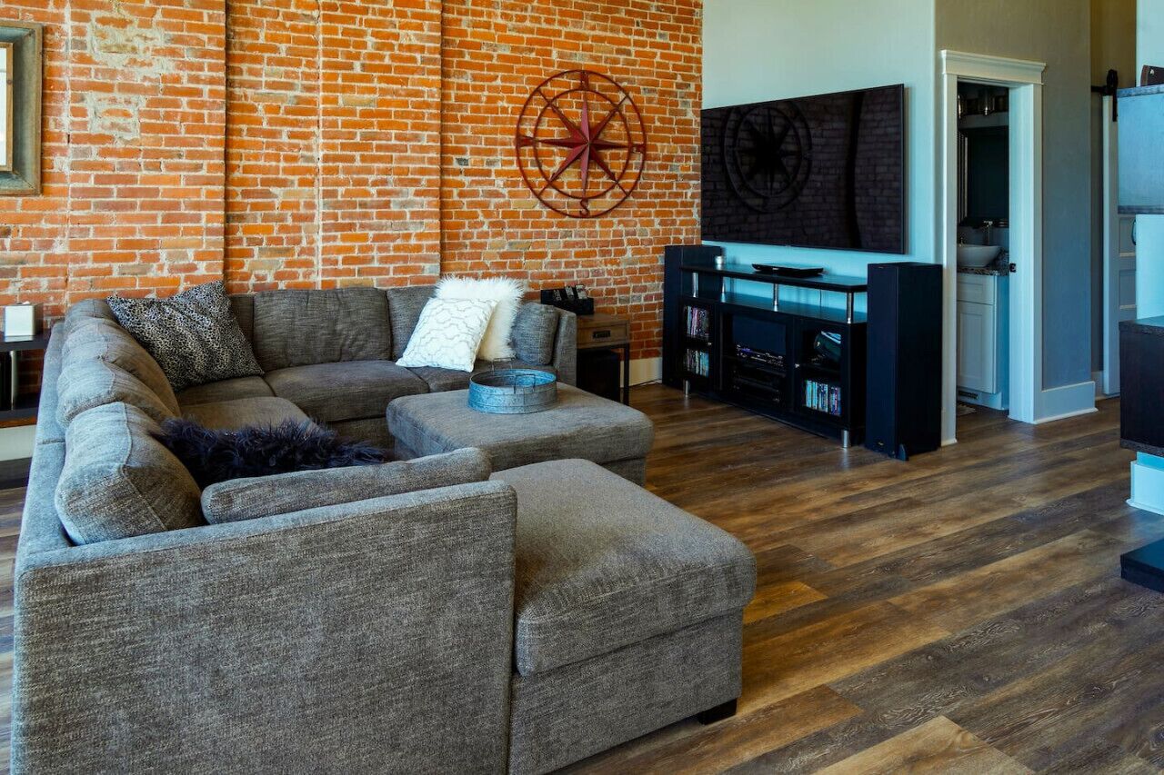 Una sala de estar con pared de ladrillo, un sofá modular grande, una mesa de centro y un mueble para televisión con televisor de pantalla plana.