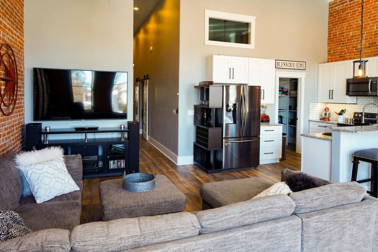 Een moderne woonruimte met een flatscreen televisie, een grote bank, een keuken met een koelkast, en een bordje met de tekst 'BLESSEUR HOME'.