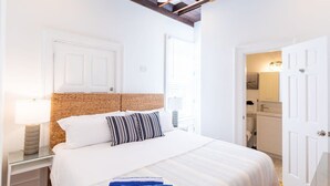 Une chambre avec un grand lit, une table de chevet avec une lampe, un plafond en bois et une salle de bain visible à travers une porte ouverte.