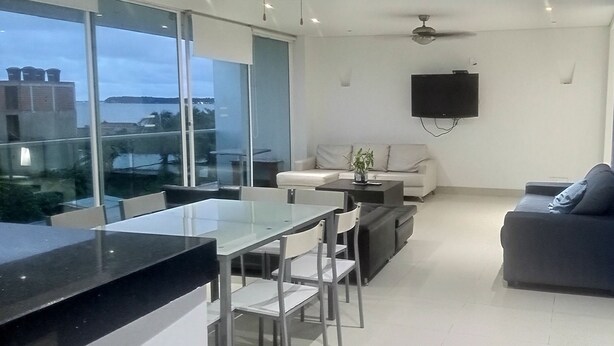A modern living room with a glass dining area, a sofa, a TV mounted on the wall, and a view of the sea. - Luxury apartment playa azul cartagena 200m² (La Boquilla)