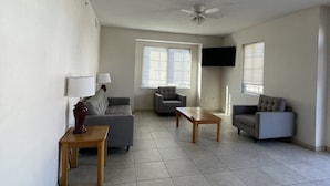A spacious living room with a ceiling fan, two grey sofas, a wooden coffee table, and a TV mounted on the wall. - Americana Princess Suites and Condos (Ocean City)