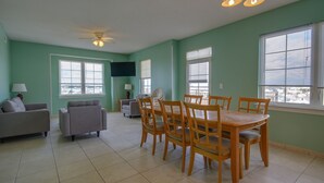 A spacious living room with a dining area, large windows, and a ceiling fan. - Americana Princess Suites and Condos (Ocean City)