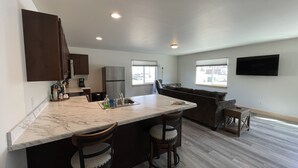 A modern kitchen with a marble countertop, two bar stools, a built-in refrigerator, and a dining area with a sofa and a TV mounted on the wall.