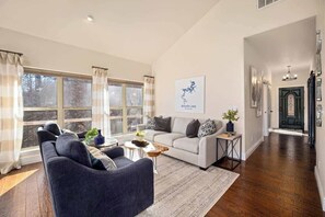 A living room with a sofa, a coffee table, and a rug. There is a window with curtains, a wall-mounted picture, and a hallway leading to a door.
