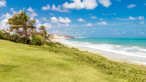 A coastal landscape with a grassy area, palm trees, and a beach. - Apartamento Novo e Decorado In Mare Bali (Parnamirim)