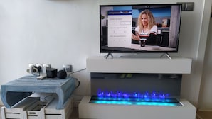 En platt-TV placerad ovanpå en vit TV-bänk, ett litet träbord med en fjärrkontroll och en blåbelyst vattenfontän under bänken.