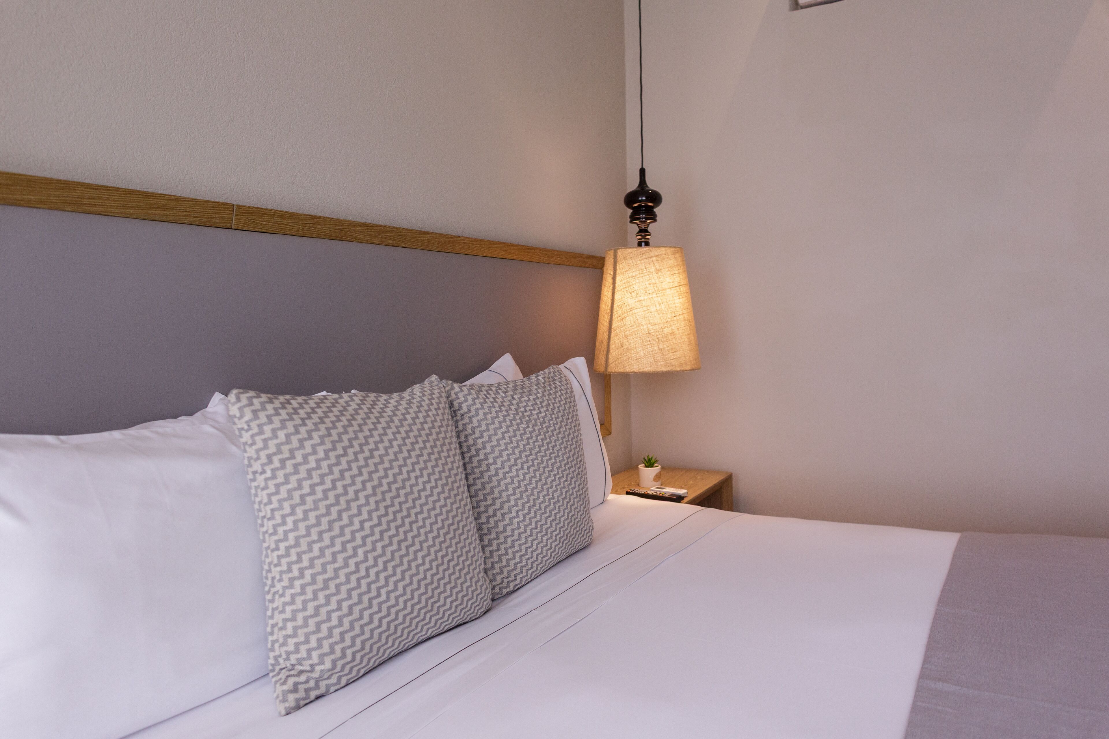 A neatly made bed with white linens and two patterned pillows, a bedside table with a lamp, and a wooden headboard.