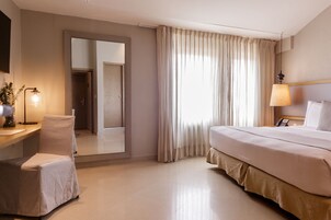 Une chambre d’hôtel avec un lit, une chaise, un bureau avec une lampe, un miroir et une fenêtre avec des rideaux.