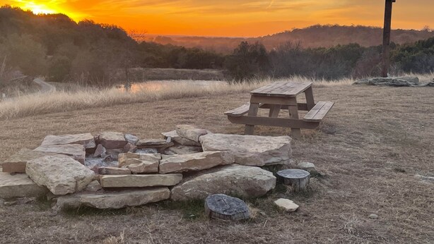 A picnic table and stone fire pit in a grassy field with a sunset in the background. - Top of the Dam Group Campground at Lake Godstone (Graford)