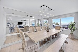 A modern dining area with a long table, wicker chairs, and a view of the ocean.