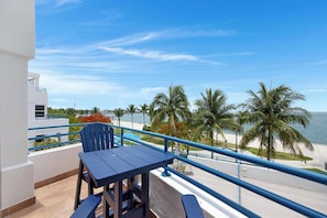 A balcony with a table and chairs, overlooking a beach with palm trees and a clear sky.