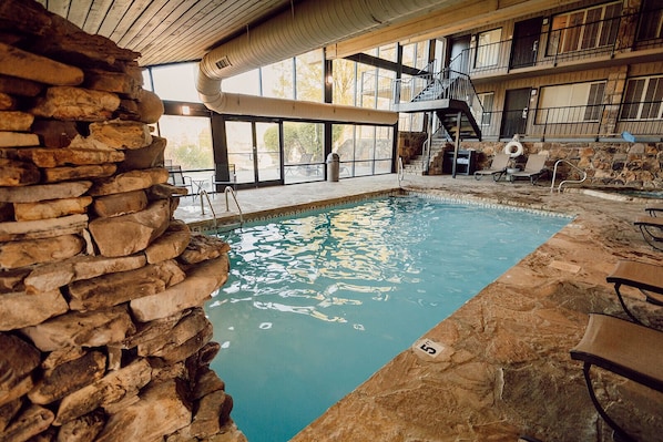 An indoor swimming pool with stone walls, a wooden ceiling, and a balcony with a staircase.