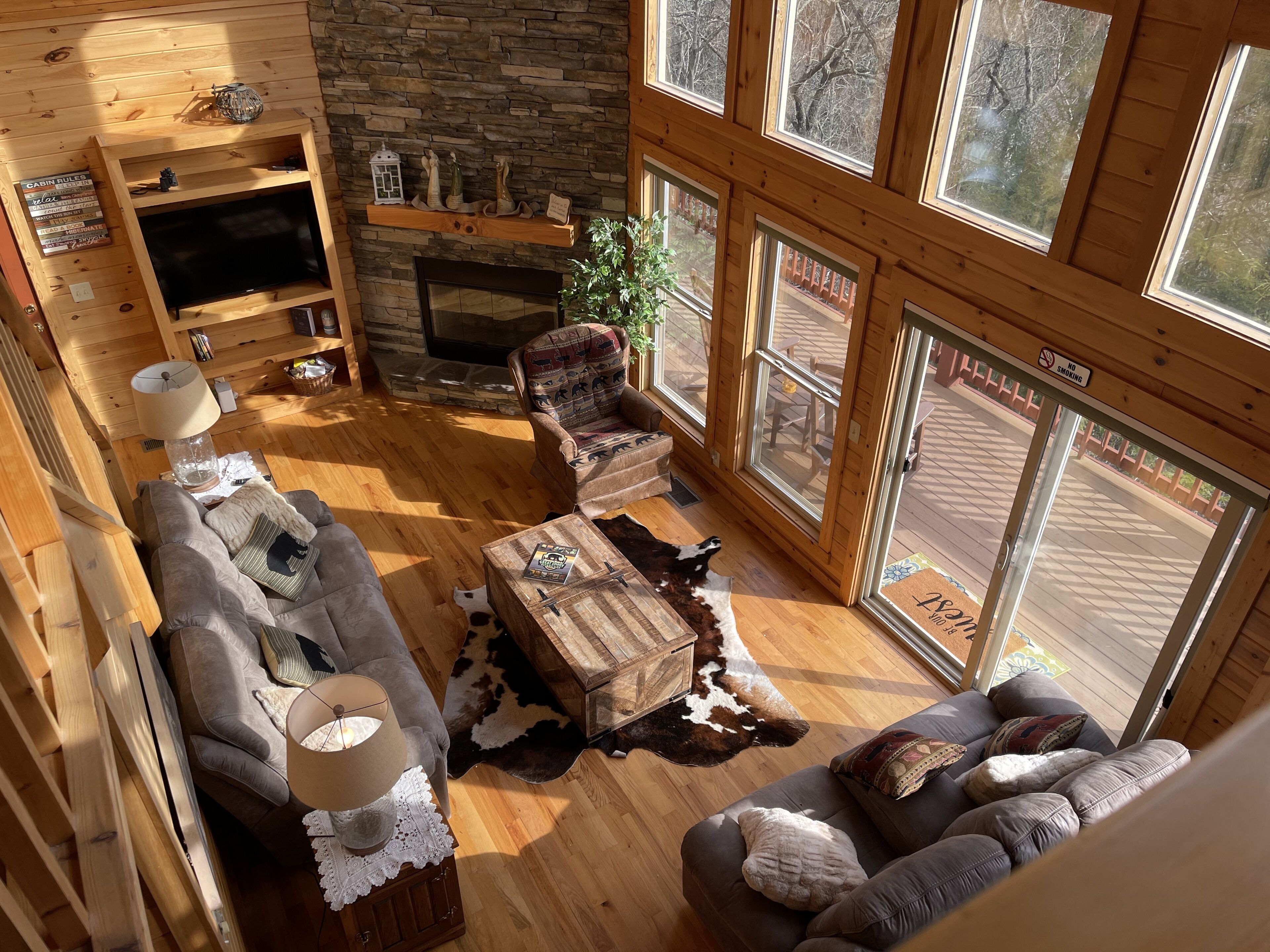 A cozy cabin interior with a stone fireplace, wooden furniture, and large windows offering a view of the outdoors.