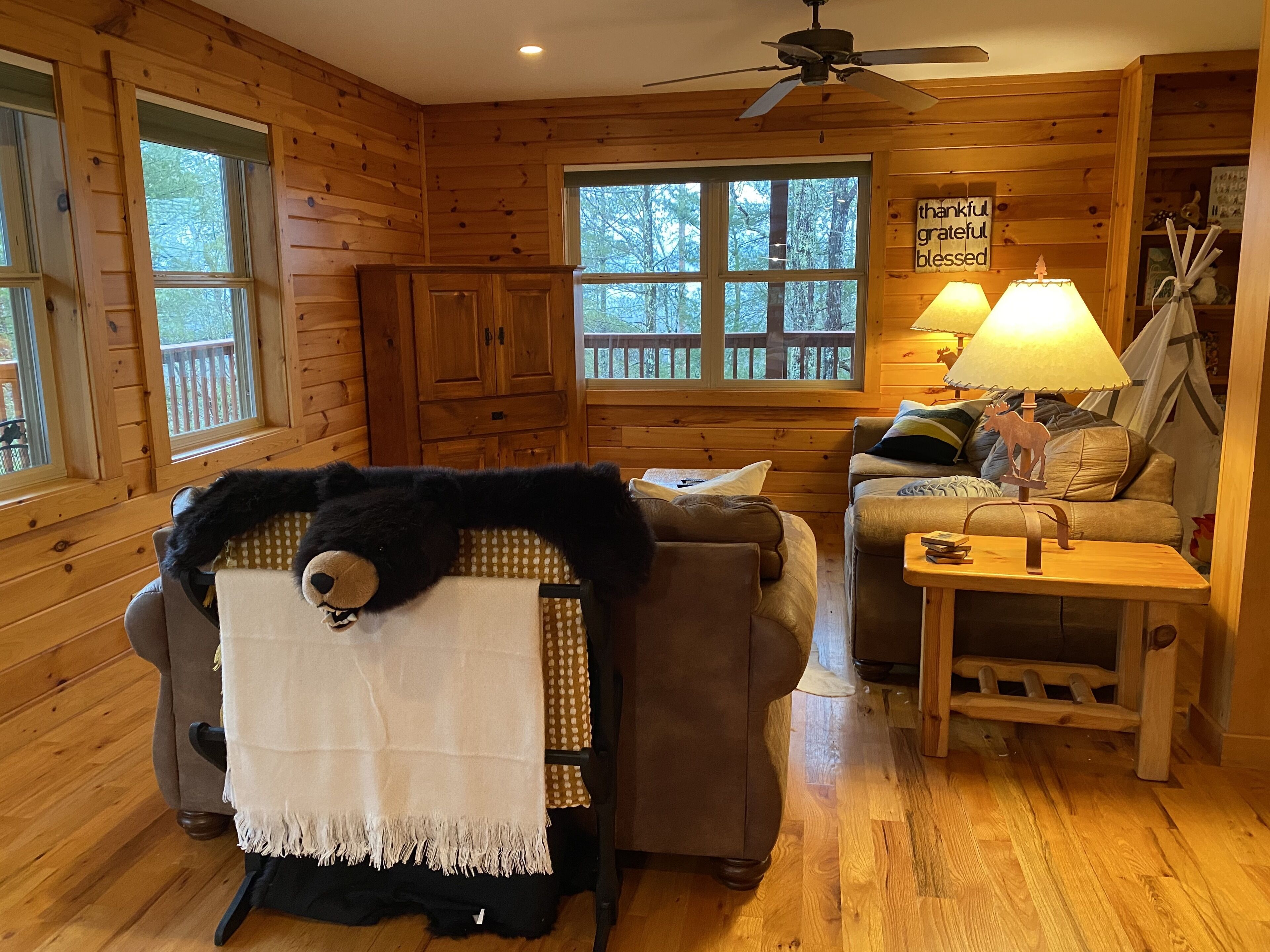 L’intérieur chaleureux d’un chalet avec des murs en bois, un ventilateur de plafond, un canapé, un fauteuil recouvert d’une couverture à motif d’ours, une lampe et une pancarte sur laquelle on peut lire : « reconnaissant, plein de gratitude, béni ».
