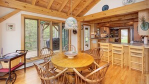 Un vaste espace dédié à la salle à manger, avec une table ronde en bois et des chaises, un lustre, et une vue sur l’extérieur grâce à de grandes fenêtres.