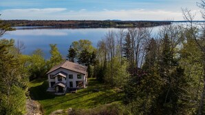 Une maison à deux étages dotée d’un grand porche à l’avant, entourée d’une zone boisée et bordée d’un lac.