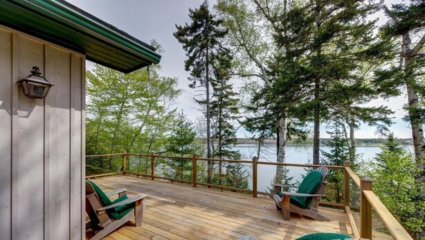 Une terrasse en bois avec deux fauteuils Adirondack verts donnant sur un lac, le tout entouré d’arbres.