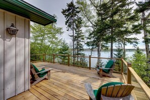 A wooden deck with two green Adirondack chairs overlooking a lake, surrounded by trees.