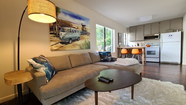 A modern living room with a sofa, a coffee table, and a painting of a van on the wall. There is a kitchen area with a refrigerator, stove, and chairs. - Coastal Zen Den (Alameda)
