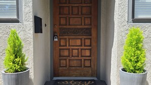A wooden door with a decorative panel, flanked by two potted plants, and a doormat with the text "story while". - Coastal Zen Den (Alameda)